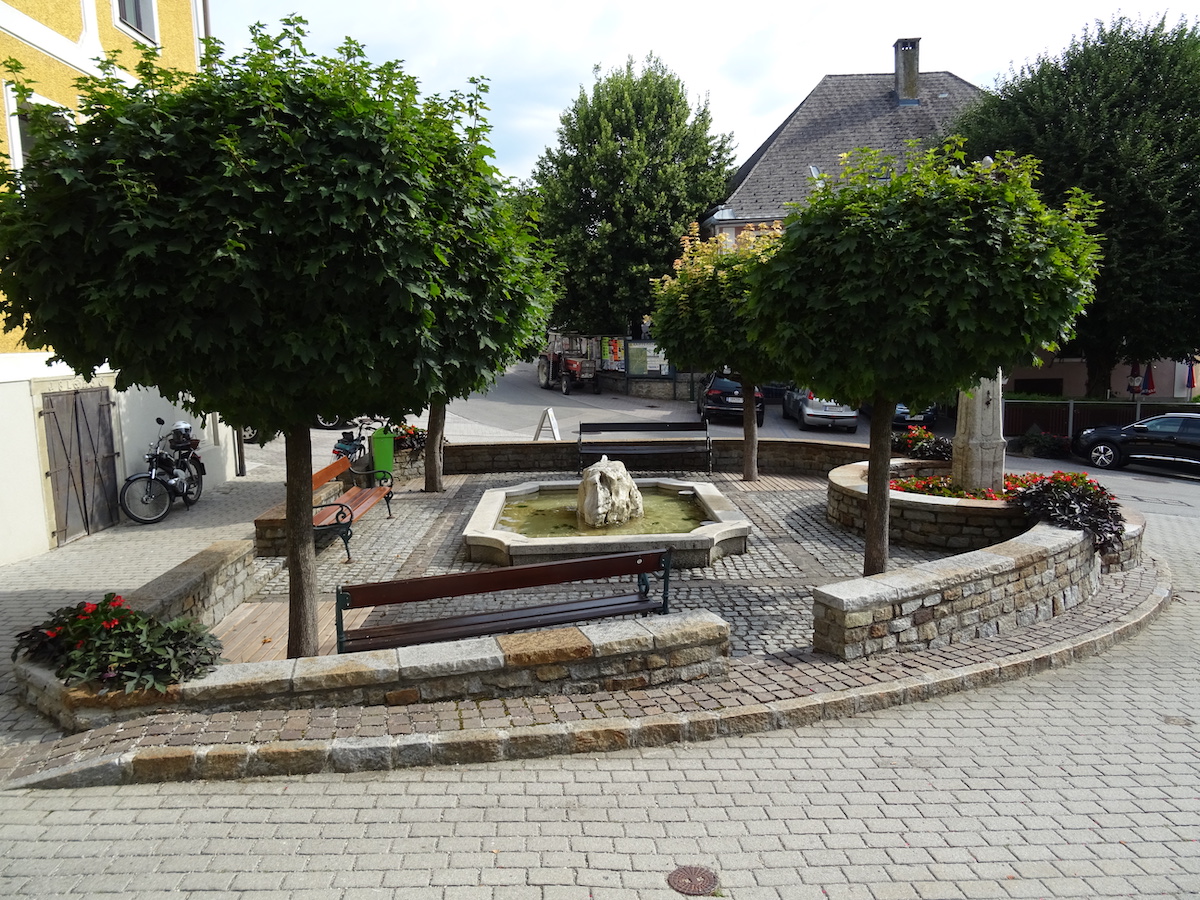 Ein kleiner, runder Steinplatz mit einem zentralen Brunnen und einer Felsformation. Bänke umgeben den Platz, gesäumt von gepflegten Bäumen und farbenfrohen Blumenbeeten. Im Hintergrund sind nahe gelegene Gebäude und geparkte Autos zu sehen.