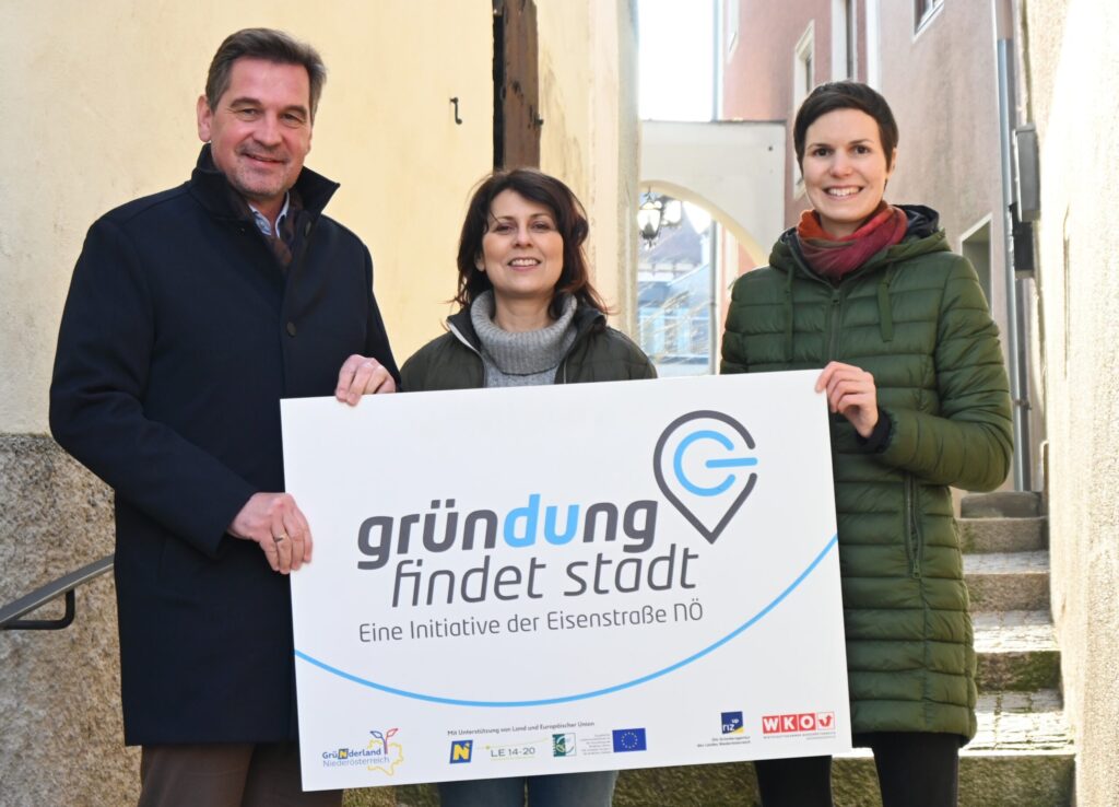 Drei Personen stehen in einer engen Straße und halten ein Schild mit der Aufschrift „Gründung findet Stadt“. Die linke Person trägt einen dunklen Mantel, die mittlere eine graue Jacke und die rechte eine grüne Jacke und einen roten Schal. Sie lächeln in die Kamera.