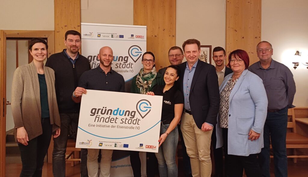 Eine Gruppe von zehn Personen steht lächelnd in einem Raum und hält ein Schild mit der Aufschrift „Gründung findet Stadt“. Sie befinden sich in einem Raum mit Holzwänden und -türen, was an eine Tagung oder Veranstaltung erinnert.