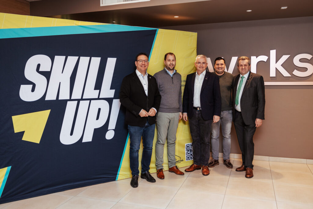 Fünf Männer stehen zusammen in einem Flur neben einem großen Schild mit der Aufschrift „Skill Up!“. Die Atmosphäre wirkt professionell. Alle tragen Business-Kleidung, die Stimmung ist positiv.
