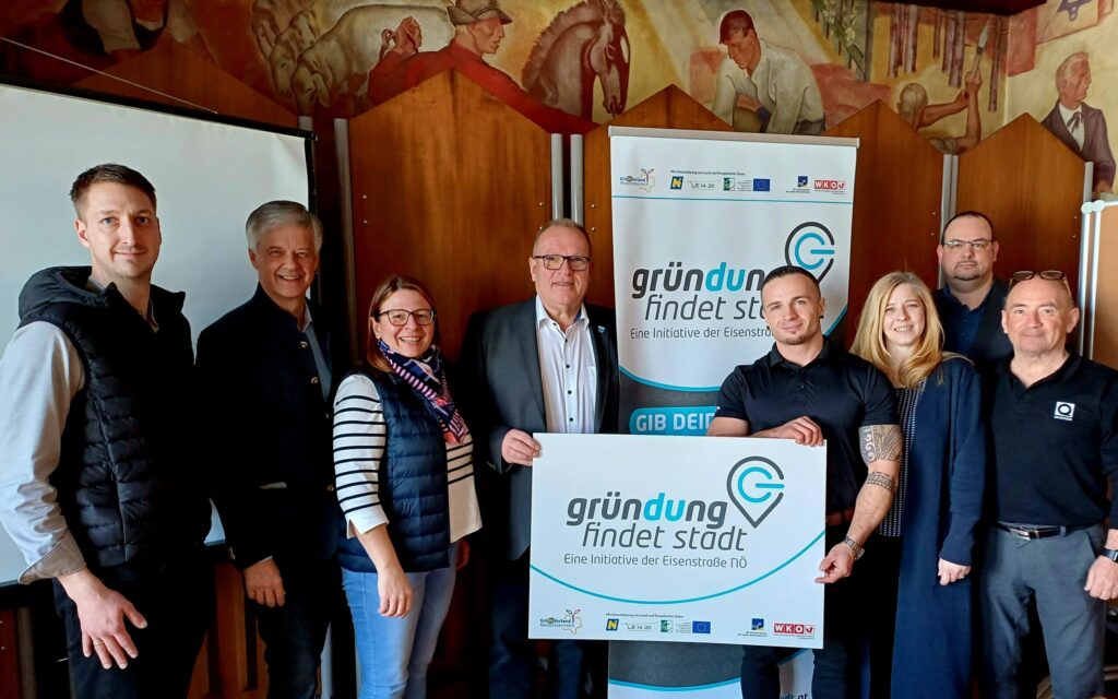 Eine Gruppe von acht Personen steht zusammen und hält ein Schild mit der Aufschrift „Gründung findet Stadt“ hoch, das auf ein Unternehmen oder eine Gemeinschaftsinitiative hinweist. Sie befinden sich in einem Gebäude vor einer hölzernen Kulisse, über der ein Kunstwerk zu sehen ist.