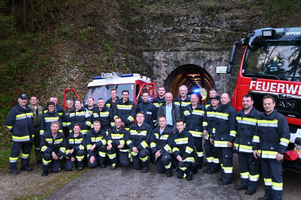 Eine Gruppe Feuerwehrleute in Uniform posiert für ein Foto vor einem Tunneleingang. Daneben stehen zwei Feuerwehrautos, ein rotes und ein weißes. Die Szene spielt in einem Waldgebiet.