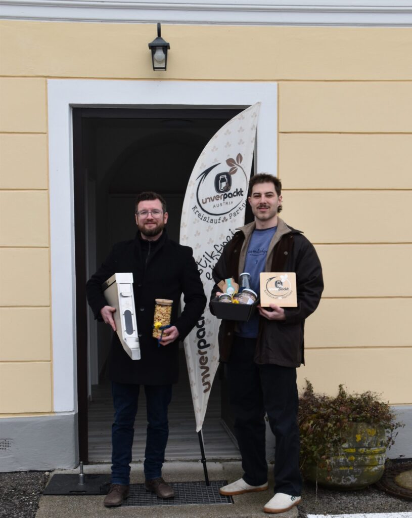 Zwei Menschen stehen lächelnd vor einem Gebäudeeingang. Einer hält eine Kaffeemaschine und ein Glas Nudeln in der Hand, der andere eine Auswahl abgepackter Lebensmittel. Zu sehen sind eine Markenflagge und Topfpflanzen.