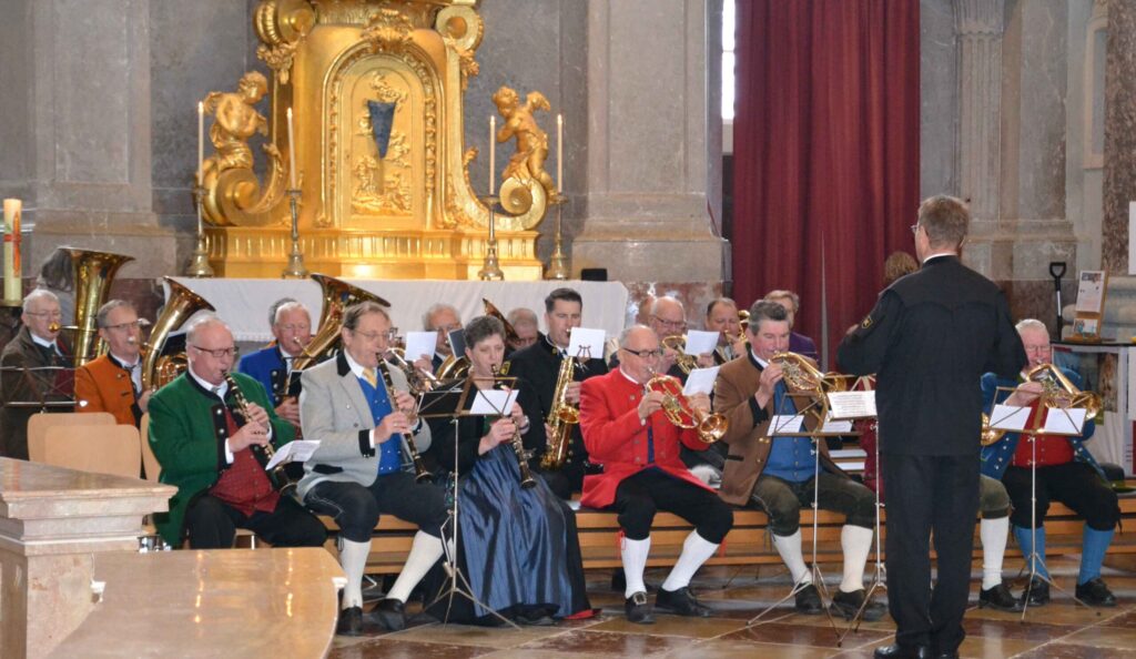 Eine Gruppe von Musikern in traditioneller Kleidung spielt in einer Kirche Blechblasinstrumente. Sie sitzen vor einem goldenen Altar, im Hintergrund ist das Publikum zu sehen. Ein Dirigent steht vor ihnen und leitet die Aufführung.