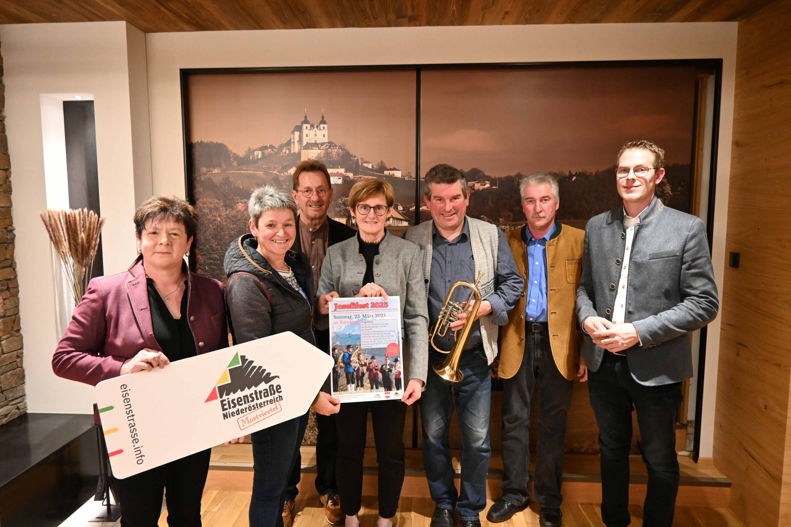 Eine Gruppe von sieben Personen steht lächelnd im Innenbereich. Eine Person hält ein Schild mit der Aufschrift „eisenstrasse.info“, eine andere eine Trompete. Außerdem hängt ein großes Plakat. Sie stehen vor einer Wand mit einem Landschaftsfoto.